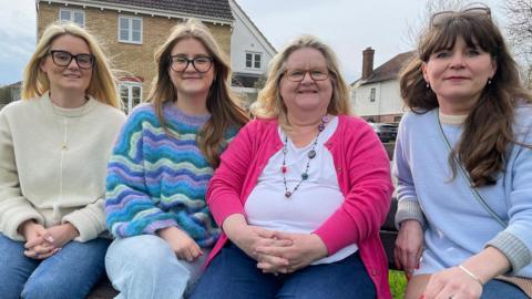 Jackie, wearing a pink cardigan and white top, sits next to her three daughters on a bench, with her house in the background 