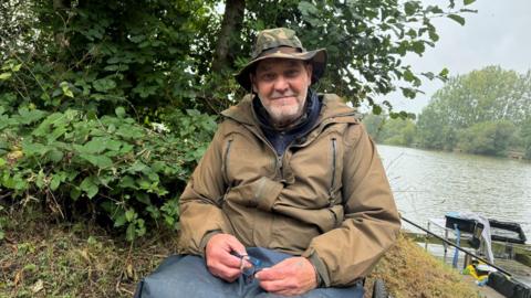 Tony Prior by the lake. He is wearing a brown jacket and holding his glasses and looking at the camera. Behind him is a tree and grass. 