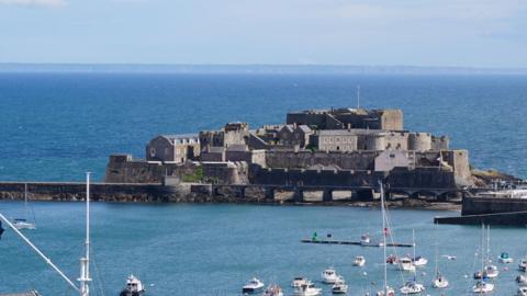 Castle Cornet from afar. A large fort structure standing inbetween St Peter Port Harbour and the open sea. 

The castle itself is brown stone and has a number of different levels, with walls that have inlets for gun emplacements. 