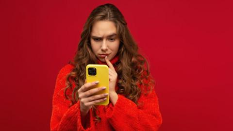 A woman in a red jumper looks quizzical as she stares at her yellow mobile phone.