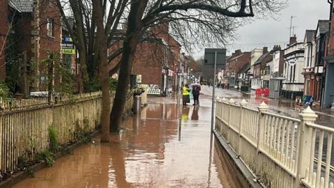 Flooding in Tenbury Wells in November, 2024. Brown floodwater on a pavement between two white fences. The street with shops in the background is also flooded. There is a small group of people in the distance. One is wearing a yellow high-visibility jacket and the other is holding an umbrella. 