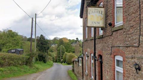 A narrow country road runs past a stone building with a hanging Castle Inn sign, surrounded by trees and hedges.