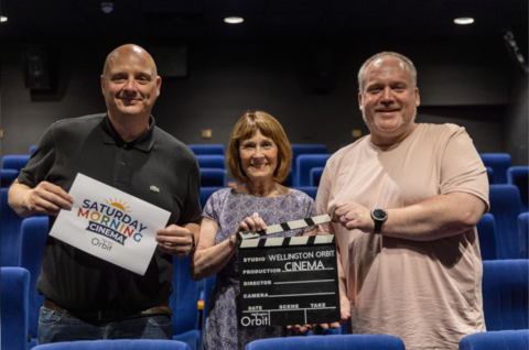 A man in a black t-shirt holds a sign saying 'Saturday Morning Cinema'. A petite lady with brown hair holds a clapperboard saying 'Wellington Orbit cinema'. A man with a neutral colour t-shirt and a black watch hold the top of the clapperboard. They are all smiling at the camera. 
