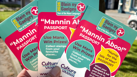 Three of the Manx passport booklets being held together. They are in tourquiose and a reddish pink colour, with "mannin aboo" in bold white lettering, and information about the Culture Vannin project.