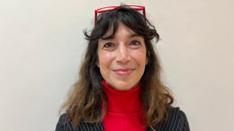 Manuela Perteghella wears a black pinstripe suit jacket with a red turtle neck collar underneath. She has red framed glasses resting on the top of her head and has long brown hair. She is smiling.