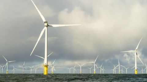 A large offshore wind farm with white turbines and yellow bases, under a cloudy sky.
