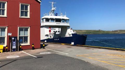 Part of the harbour area in Lerwick. A small ferry is stationary, next to a red building with cream windows. The sea and sky are blue and there's a green island in the distance. 