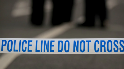 Blue and white tape carrying the instruction "police line do not cross" in capital letters. Out of focus in the distance are the legs of two or three people who are wearing dark trousers.