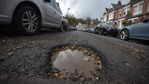 A pot hole filled with rain water and pebbles is in the foreground of a grey road on a suburban street with parked cars on either side of the road outside brick houses.