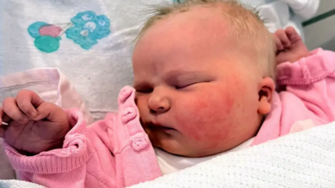 A close up photo of a baby, lying on her back with her head to the side and eyes closed, wearing a pink cardigan and with a white blanket reaching just below her chin.