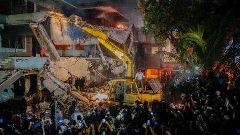 A crowd in front of a burning building, with an excavator striking the house's wall