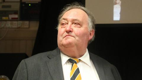 A head-and-shoulders shot of a man wearing a shirt, a yellow and black tie and a grey suit jacket. He has grey hair and is looking away from the camera.
