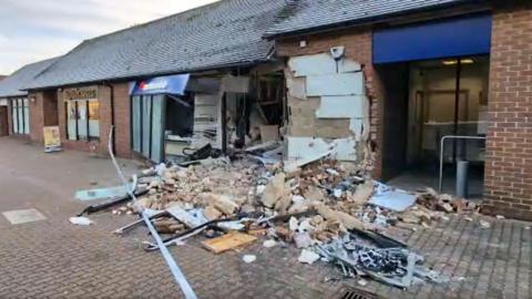 The front of a building society that has been severely damaged, showing ripped out walls, bricks and debris on the ground and an open shop front. There is police tape across the front. 
