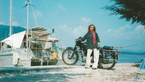 A woman leans against her motorbike next to a harbour. The skies are blue and sunny and there is a white yacht to the left of the woman and her bike.