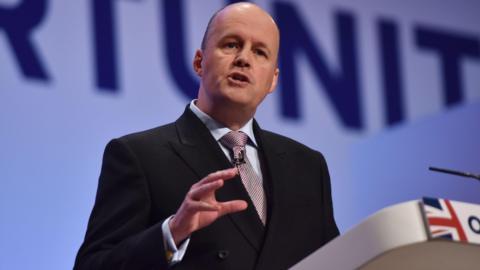 A photo taken at a conference with Sir Ashley fox wearing a suit, speaking at a lectern with one of his arms raised slightly. 