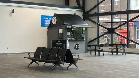 Caribe Coffee. The small hut has been built in the corner of Sunderland Station. Two tables with four chairs are set out of the front of the hut. The rest of the station is empty.