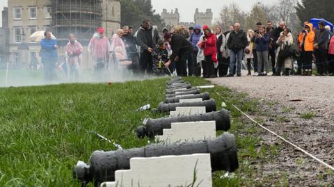 People stood in the rain with mini cannons laid out in front of them on a lawn.