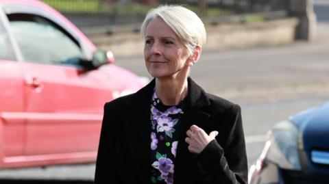 Lady Eleanor Donaldson has short blonde hair. She is wearing a black blazer and a floral blouse. Behind her a red car can be seen.