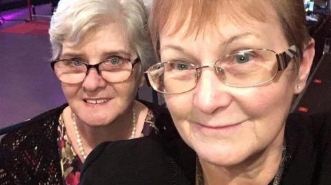 Two women pictured together, both are wearing glasses and have short hair. The woman on the left has a white necklace on and black shawl. The woman on the right wears a black jacket. 