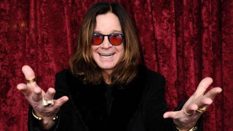 A man with long brown hair and red sunglasses with his hands outstretched and a black top in front of a red curtain
