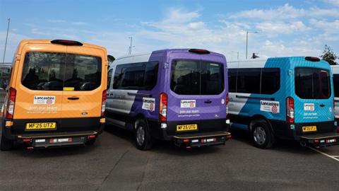 The backs of three of the minibuses parked up on a car park under a blue sky, one is orange, the middle one purple and the one on the right is turquoise