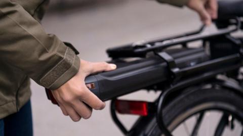 A close-up of someone's hand on a long black battery that they are fitting on to an e-bike. The bike is black and the person is wearing a green coat, you can't see anything above where their arms are.