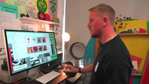 A man with blond hair and a black sweatshirt looks at designs on his computer