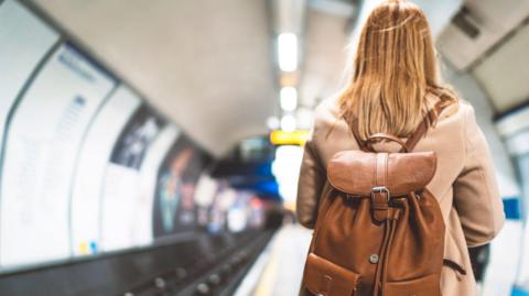 A woman with long blonde hair wearing a light brown coat and a brown leather backpack stands with her back to us on a London tube platform. 