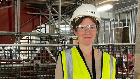 Project manager Verity Bennett stands in front of a blue barrier wearing yellow high-vis, a white hard hat and pink gloves. Behind her is a scaffold.