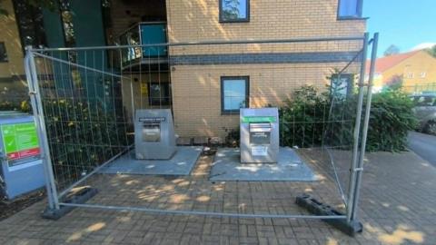 Two large silver metal rubbish chutes, with a large metal fence around them. Behind them is a block of flats, there are some small hedges either side of the bins.