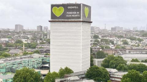 Grenfell tower which is covered in white hoardings and a green and grey sign around the top floors saying "GRENFELL FOREVER IN OUR HEARTS" and a green heart. 