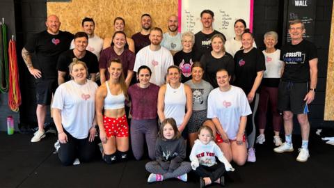 A group of 22 people posing for a photo in a CrossFit gym. They are all wearing gym gear and standing or kneeling in three rows, smiling at the camera. 