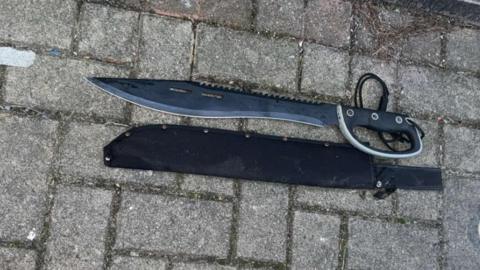 A large knife with a dark blade lies on the ground next to a wall. The blade is sharpened on one side, with serrations on the other and is attached to a hilt with a hand guard.
