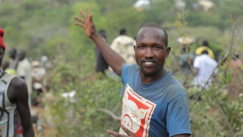 Manasse Lomachar in a blue T-shirt with a cartoon pig printed on it looks at the camera but gestures behind him with his right hand. In the background, out of focus, people can be seen working among the shrubs.