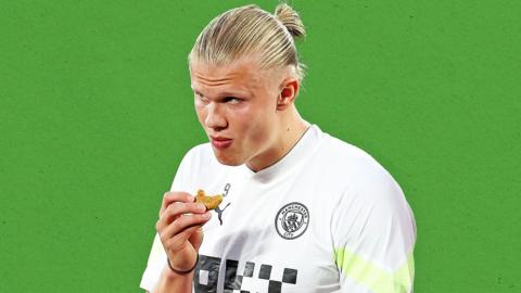Erling Haaland looks up as eats a snack. His blonde hair is tied back