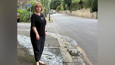 A woman wearing black trousers and a top standing on a pavement next to a street. The picture shows a damp area where water has been flowing.