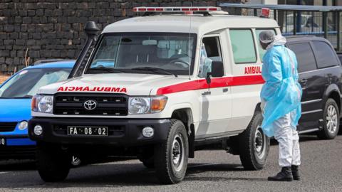 A truck-style ambulance is stopped on a street while a person in a hazmat suit stands next to it talking to the driver, also in a hazmat suit, as they evacuate a person from the ship to the airport to be transferred to the Netherlands for treatment, in Cape Verde on Wednesday.