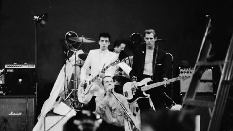 The Clash in black and white. One member of the band is on his knees singing into a microphone while other members of the band play their instruments with stern expressions behind him. They are surrounded by music amps.