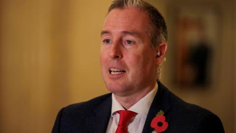 A photo of Paul Givan mid-speech at Stormont. He has short, greying hair and is wearing a black suit jacket, a white shirt, a bright red tie and a red poppy on hs lapel.