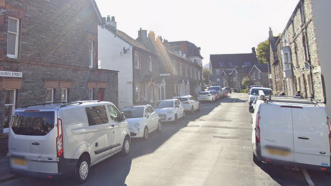 Church Street in Keswick is narrow and surrounded by stone and brick terraced houses. There are lines of parked cars on either side of the road. 