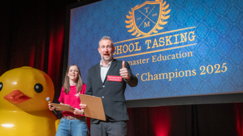 Comedian Alex Horne stands on a stage and looks at the camera doing a thumbs up. Next to him is a woman in a red shirt and blue jeans. On a screen behind them it says School Tasking and Champions 2025, although more text is obscured by Alex Horne. A very large yellow rubber duck is to the left of the picture.