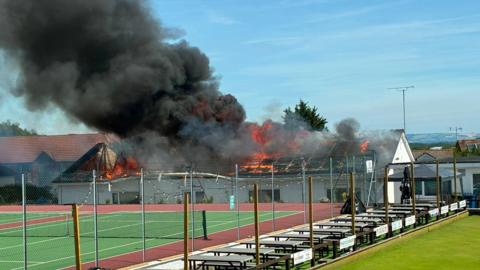 Smoke and flames billow from the roof of a social club. There is a tennis court and a bowling green in the foreground and the club is surrounded by houses. The sky is blue and there is some seating between the bowling green and the tennis court.
