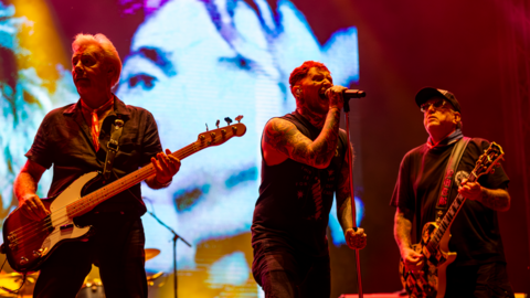Members of the Sex Pistols playing on stage; the man in the middle is singing into a microphone flanked by two men playing guitars