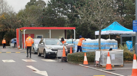 A car stopped next to people in high vis jackets and pallets of bottled water. It is in the car park of tesco in Henley.