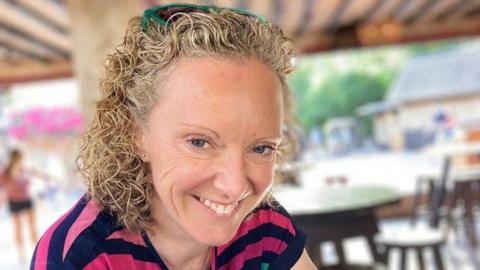 A woman with mid-length blonde curly hair smiles at the camera. She wears a pink and navy striped top and has green sunglasses on her head.