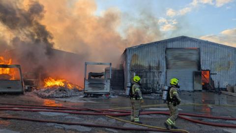 Firefighters in an industrial site covered in water stand together, wearing protective gear, while several orange fires blaze nearby. Smoke is rising to cover the blue sky.