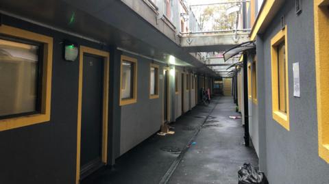 A passageway between two rows of two-storey flats with yellow rimmed windows and grey walls. There is a black plastic rubbish bag in the foreground and yellow steps at the end of the passageway. There are connecting walkways on the first floors between the two rows.