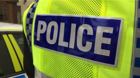 The back view of a yellow British police vest with the words police in white block lettering on a blue horizontal strip.