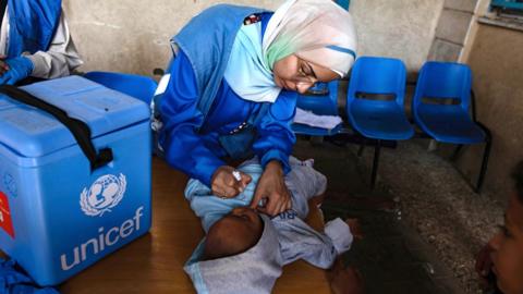 A small Palestinian child lies on a table and receives oral polio vaccine drops from a healthcare worker during a vaccination campaign at UNRWA headquarters in Deir al-Balah, central Gaza Strip, 14 October 2024 