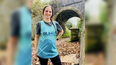 Christine Lawrence in a turquoise running top with the RUH logo on it, standing smiling to camera on a track with a bridge in the background